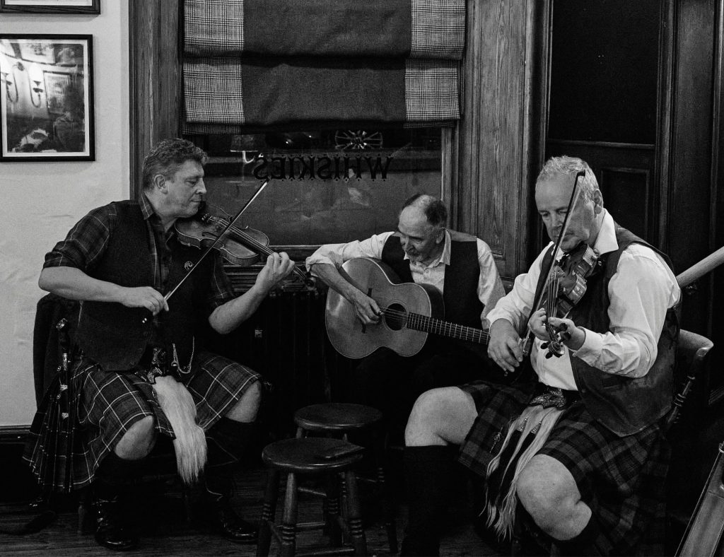 Three male Scottish folk musicians sat playing their string instruments in a pub.