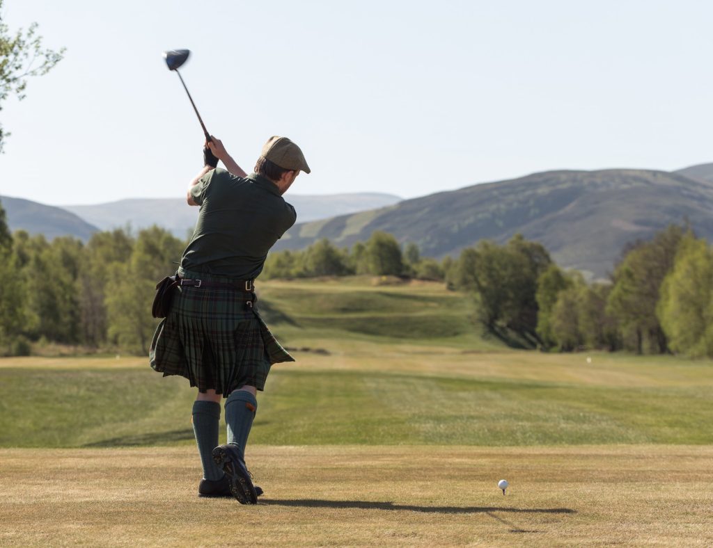 view of a golfer from behind on a sunny day teeing off and wearing a kilt.