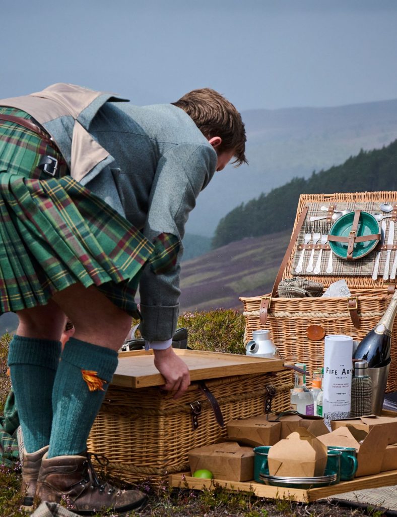 A ghillie wearing a green kilt bending over to unpack a luxury picnic hamper on top of a Scottish mountainside.