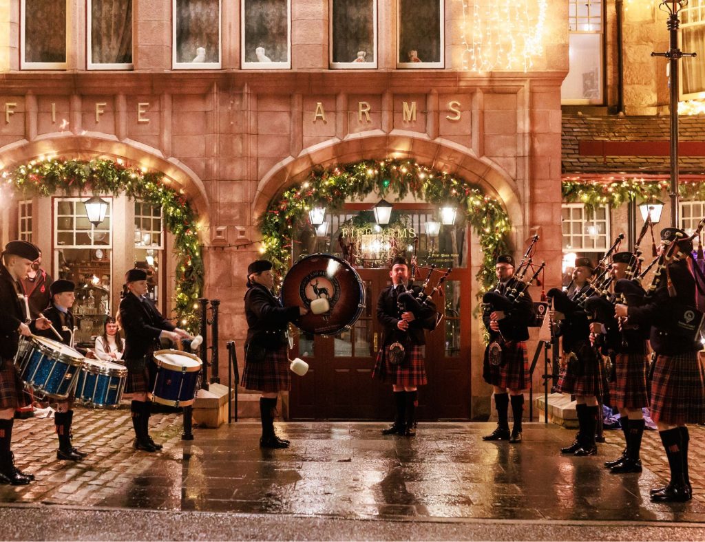 A full Scottish pipe band playing at night outside of the Fife Arms hotel lit up with fairy lights.