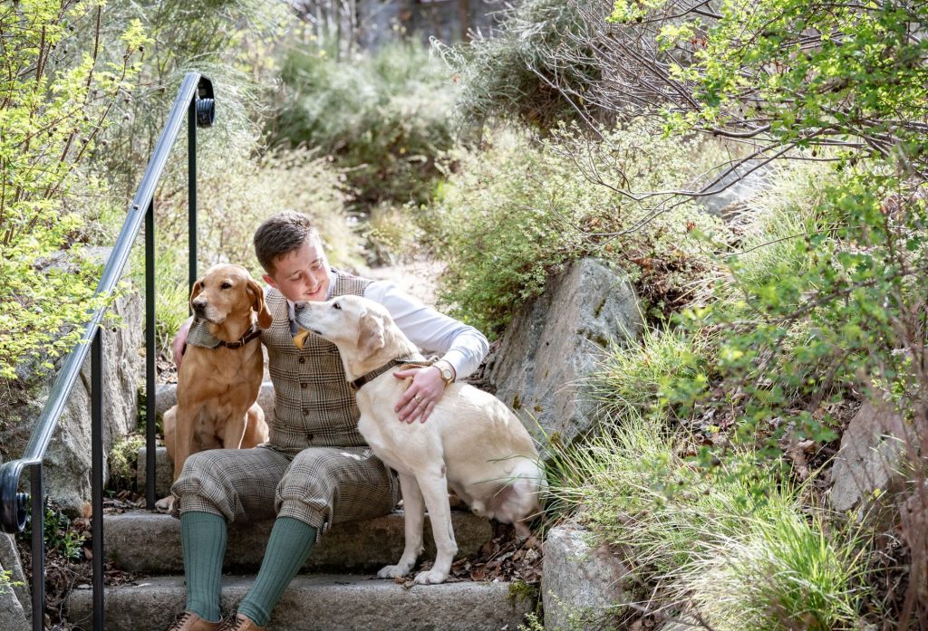 Man in tweed uniform sitting on garden steps with two Labradors.