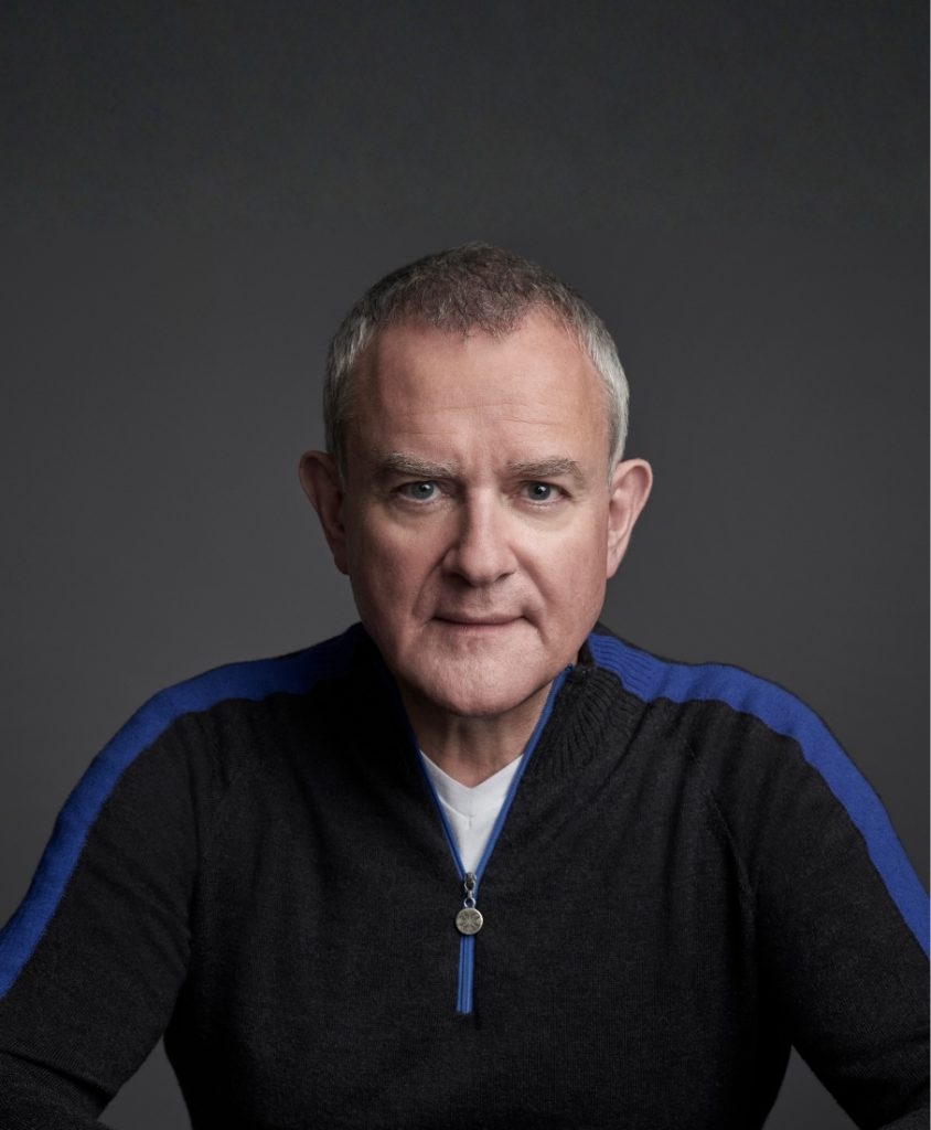 A man with clasped hands wearing a blue and black jumper.