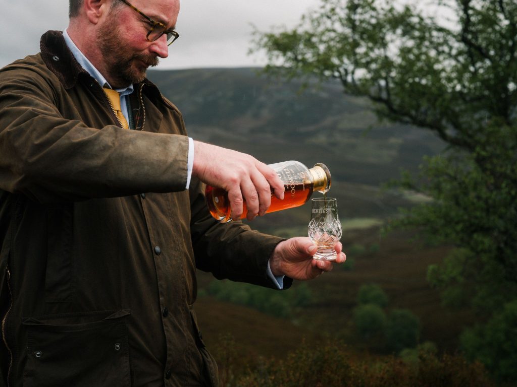 A man pouring whisky into a glass in a Scottish landscape