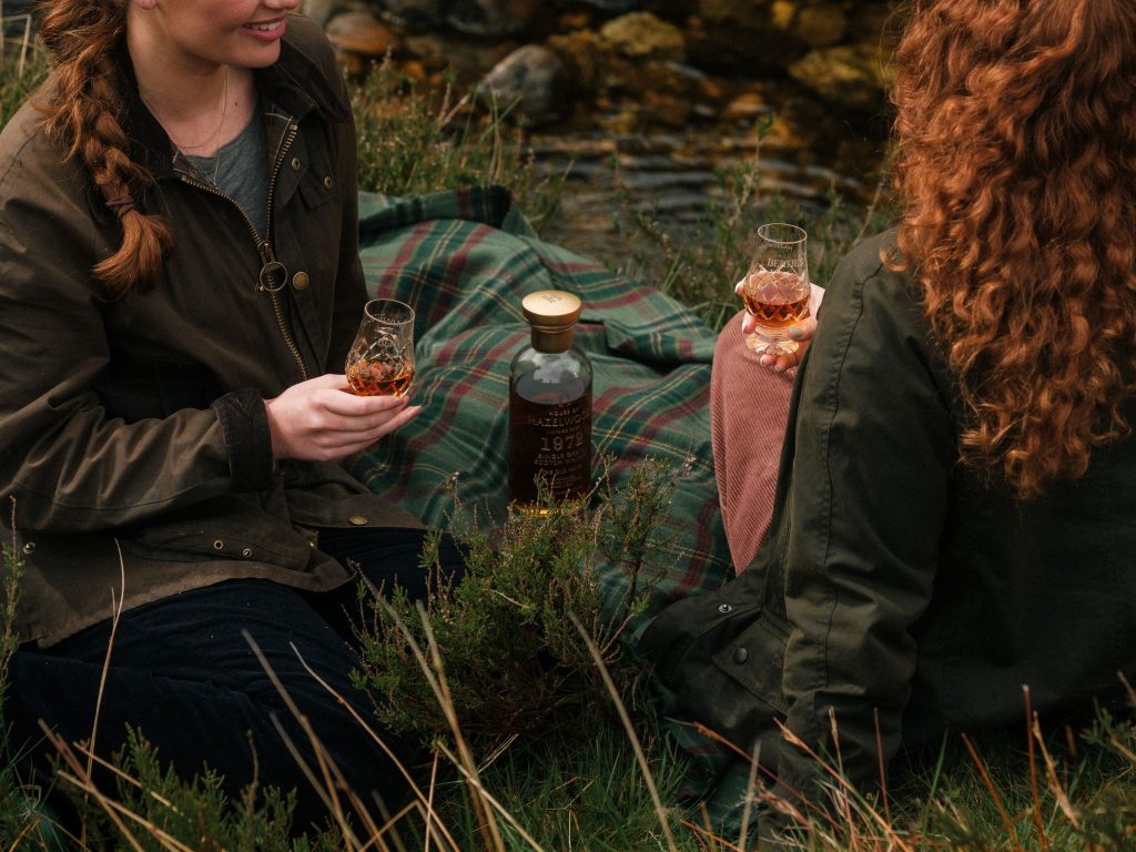 A tartan picnic blanket with two women tasting whisky