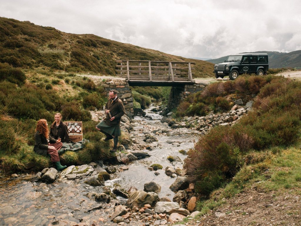 Three people tasting whisky beside a river with a Land Rover in the background