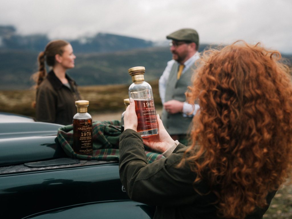 A woman with red curly hair holding a bottle of whisky with two people in the background