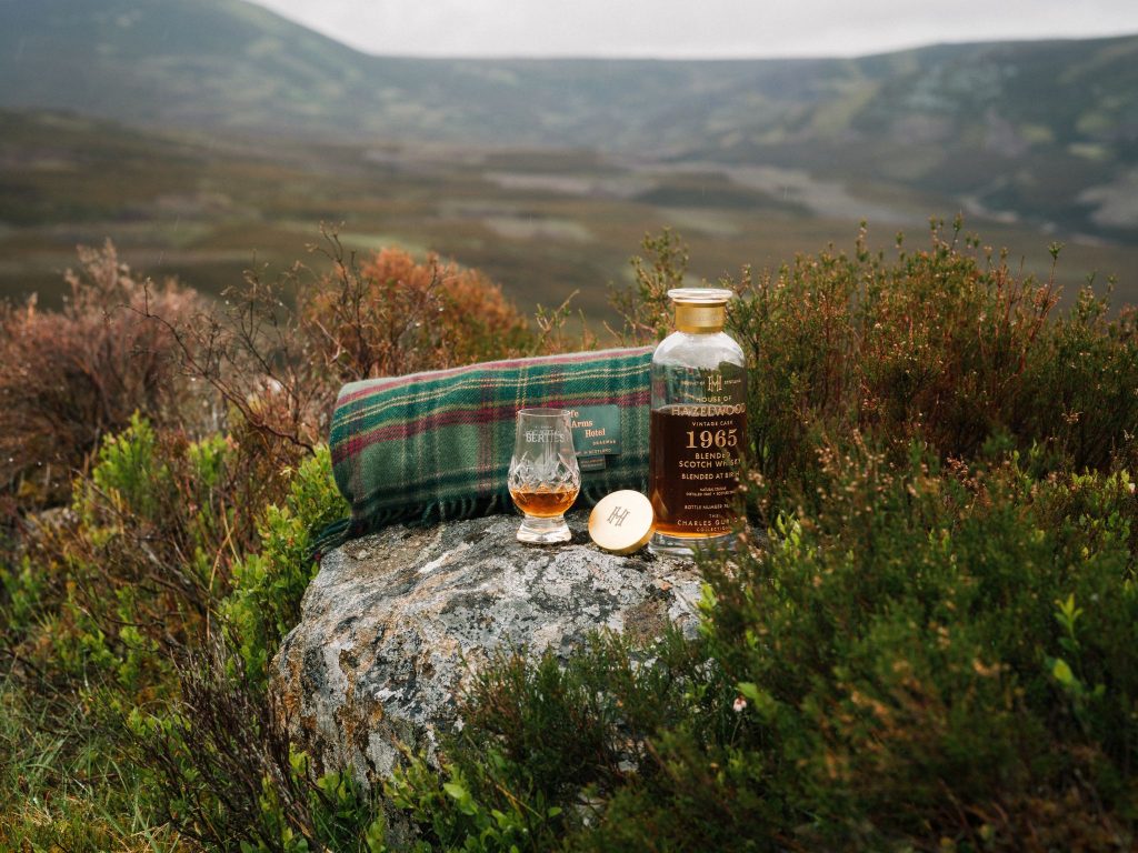 A glass of whisky, a whisky bottle, and a tartan blanket in a Scottish landscape