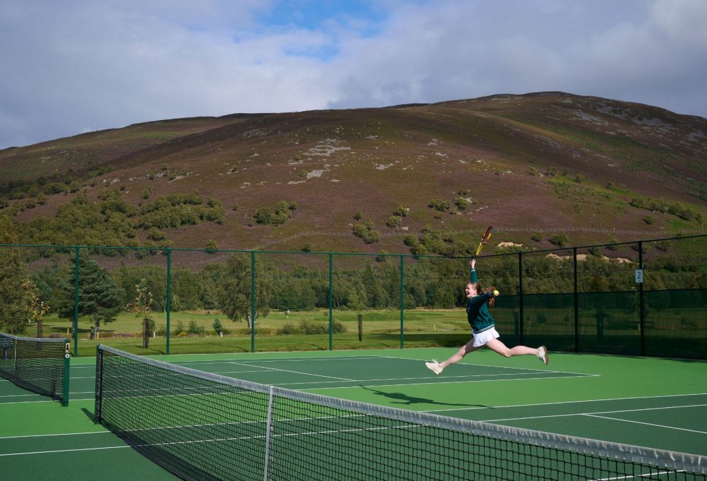 Young girl jumping in mid-air with a tennis racket and a ball on Braemar tennis court.