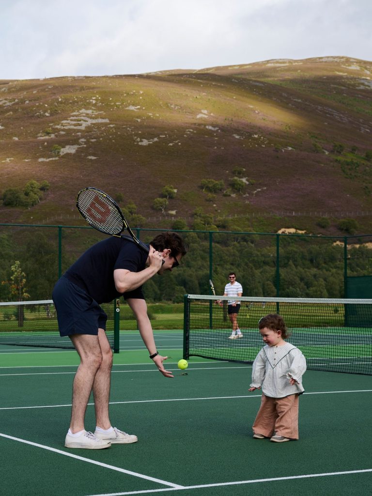 Man practicing tennis on a tennis court with a very young child.