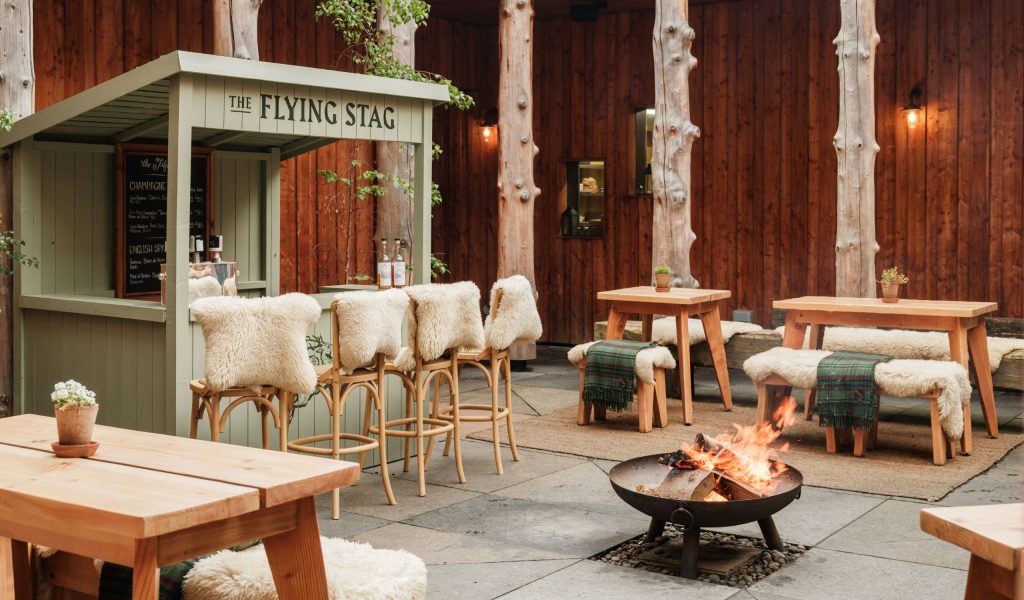 A fire pit and oyster and champagne hut surrounded by wooden seating in a courtyard