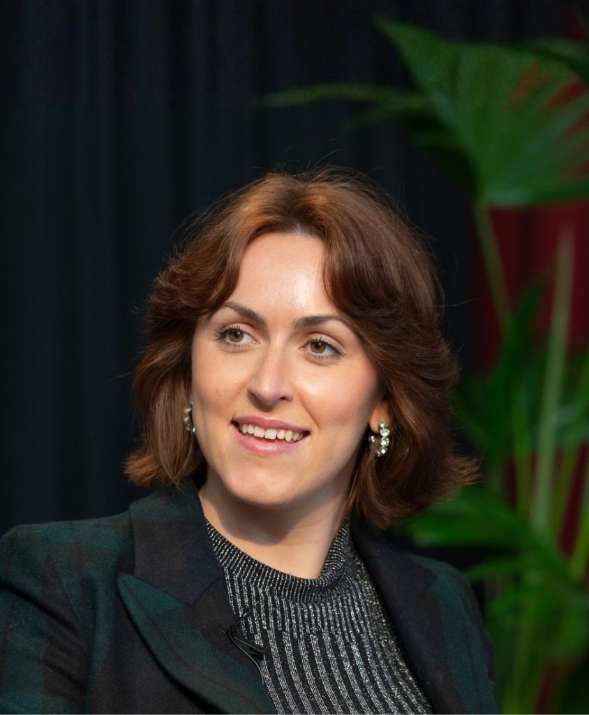 A smiling woman with brown hair and plants in the background