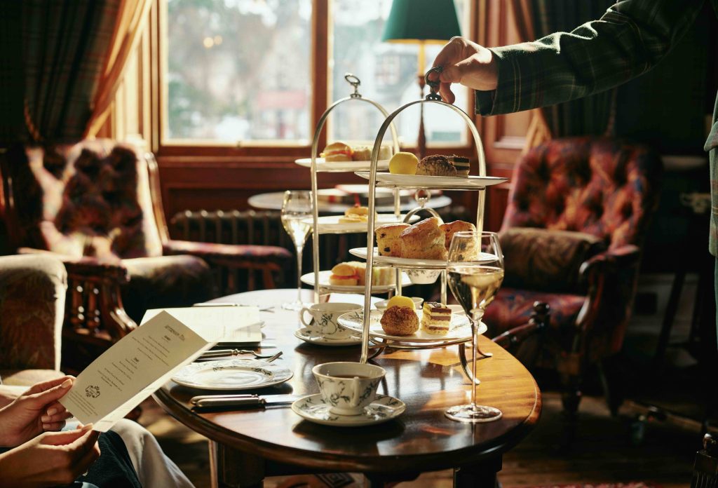 A cake stand full of sandwiches and cakes being set down on a wooden table in a luxuriously furnished drawing room, a window in the background is casting golden light on the scene.