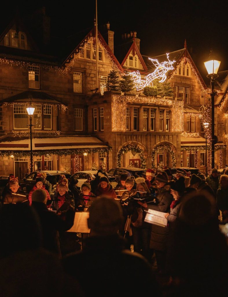The Fife Arms Hotel at night from the outside, decorated with Christmas lights with a crowd of carol singers in a circle underneath singing.