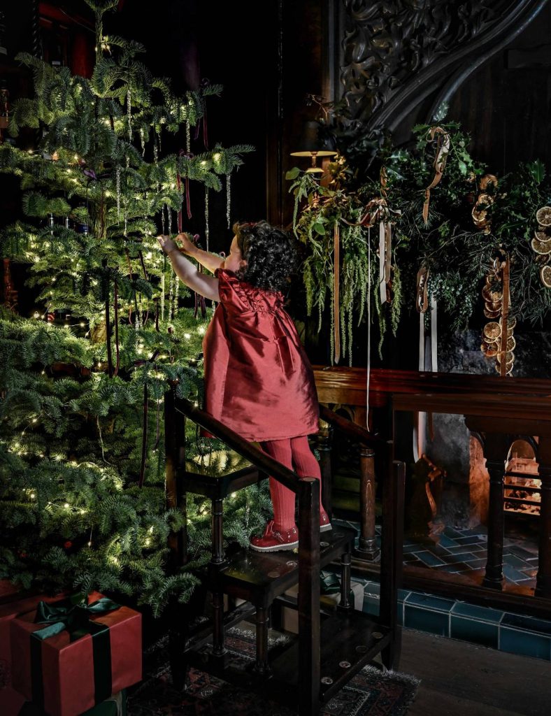 A young child in a red dress hanging a decoration on a Christmas tree laden with lights and with presents wrapped underneath.