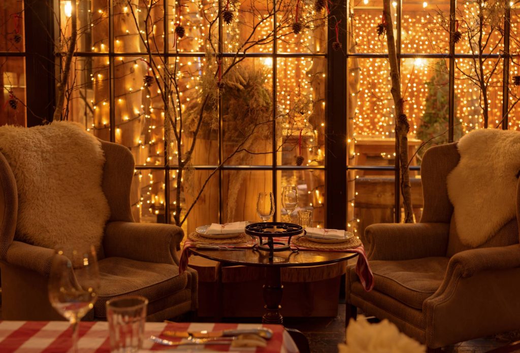 Two large armchairs to their backs to a festively lit courtyard. Each chair has a sheepskin rug on the back of it and there is a table positioned between them.