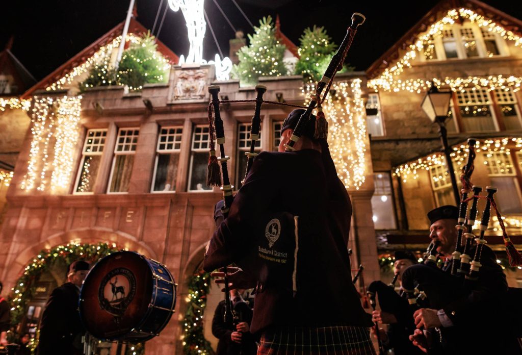 A bagpipe player outside of the Fife Arms Hotel at night, the hotel is decorated in festive lights.