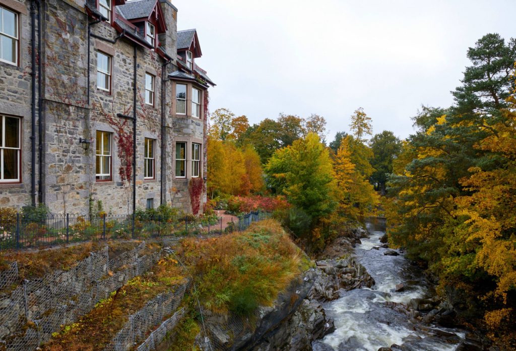 A view of the side of the Fife Arms Hotel on the left covered in red ivy, in front is the River Dee and either side are trees in autumnal colours.