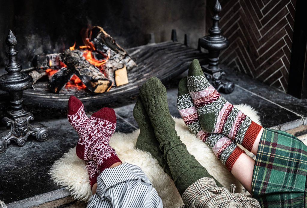 Three people's feet in front of a lit fire, each person has festive patterned or coloured, cable knit socks on.