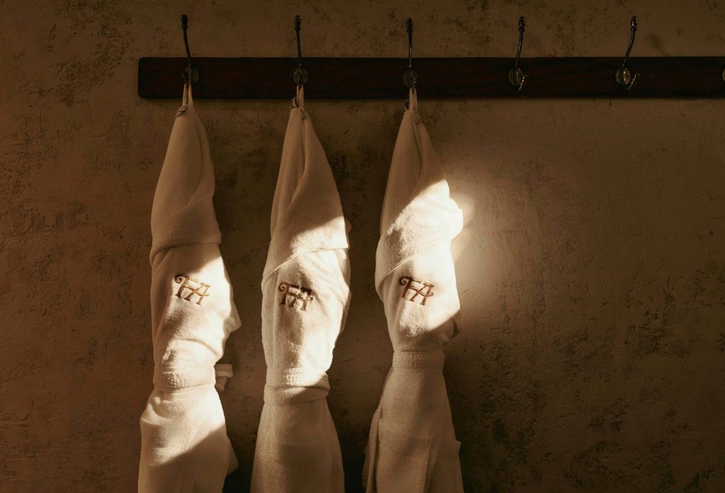Three white spa robes hanging on pegs in the sun, each is embroidered with the Fife Arms Hotel logo.