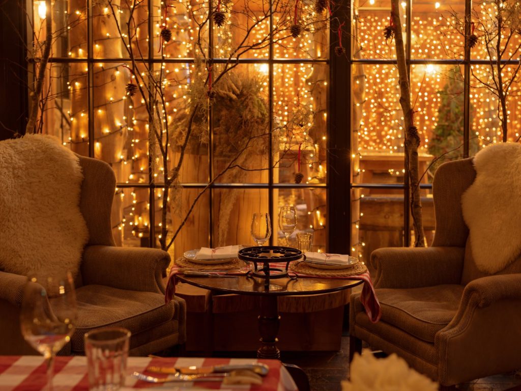 Two armchairs either side of a table in a warmly lit room, with a courtyard with fairy lights through a window in the background