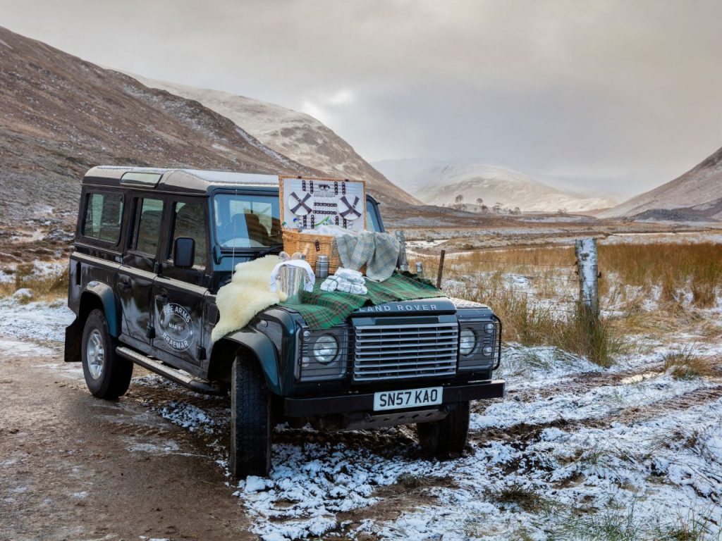 A Land Rover with a picnic basket and blanket on its bonnet in a snowy mountain landscape