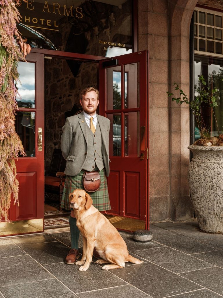 A man in traditional Scottish clothing with a golden Labrador dog