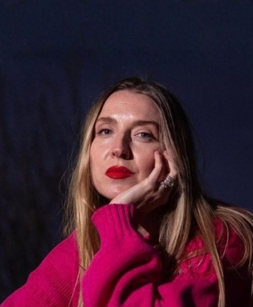A blonde woman resting her chin on her hand in a fuchsia jumper