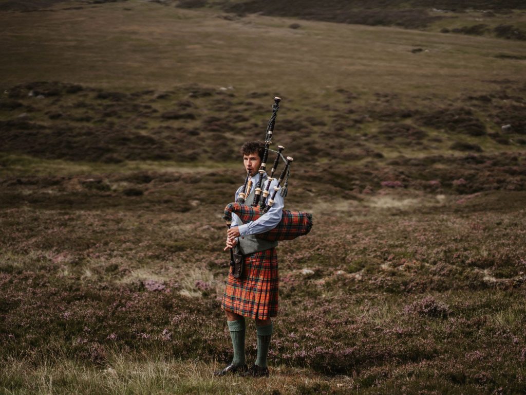 A piper playing in traditional Scottish dress in a heather moorland