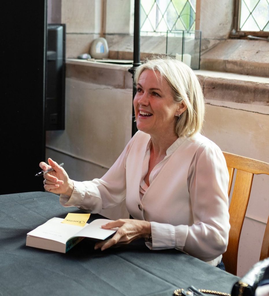A woman with short blonde hair seated and signing a book